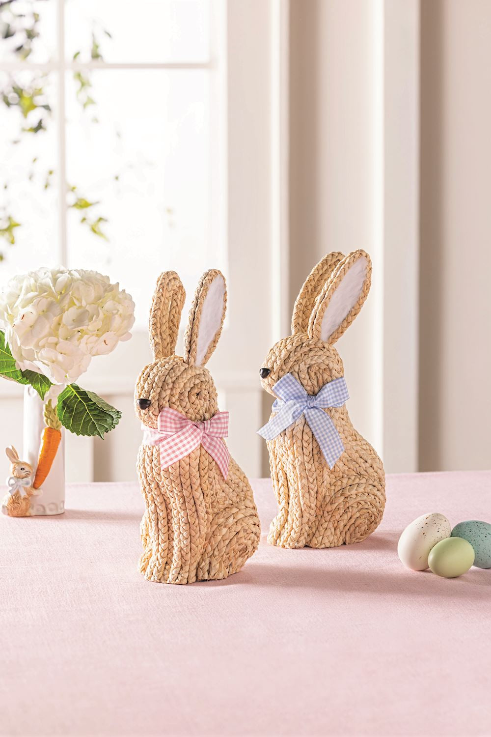 Two woven bunny figures with decorative bows on a pink surface with Easter eggs and flowers.