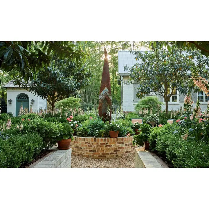 A landscaped garden courtyard with a central rusted metal sculpture rising above a brick planter bed, surrounded by dense green shrubs, potted flowers, gravel, and trees. A white house is visible in the background behind the foliage.