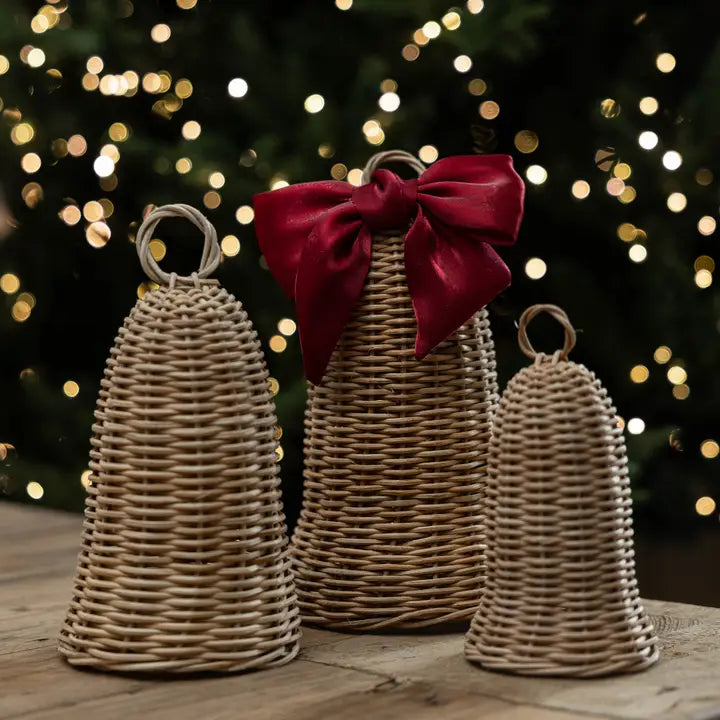 Natural woven rattan decorative bells in three sizes displayed in front of Christmas lights, with a burgundy velvet bow on the large bell.