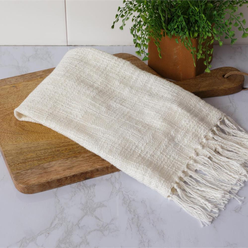 White woven blanket with fringe on a wooden cutting board, with a plant in the background.
