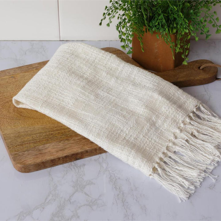 White woven blanket with fringe on a wooden cutting board, with a plant in the background.