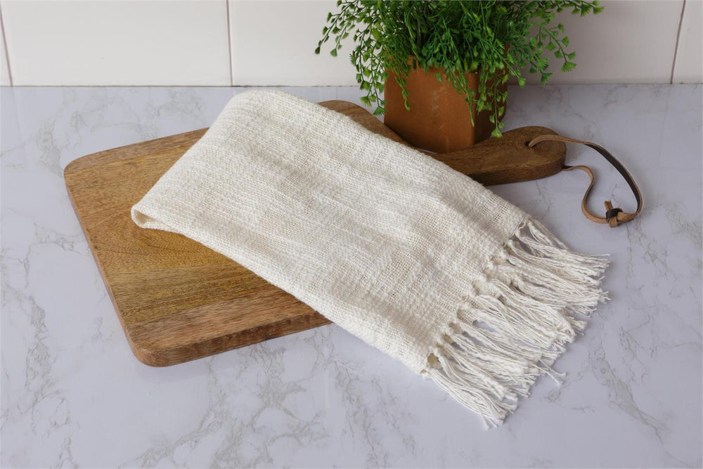White woven blanket with fringe on a wooden cutting board, with a plant in the background.