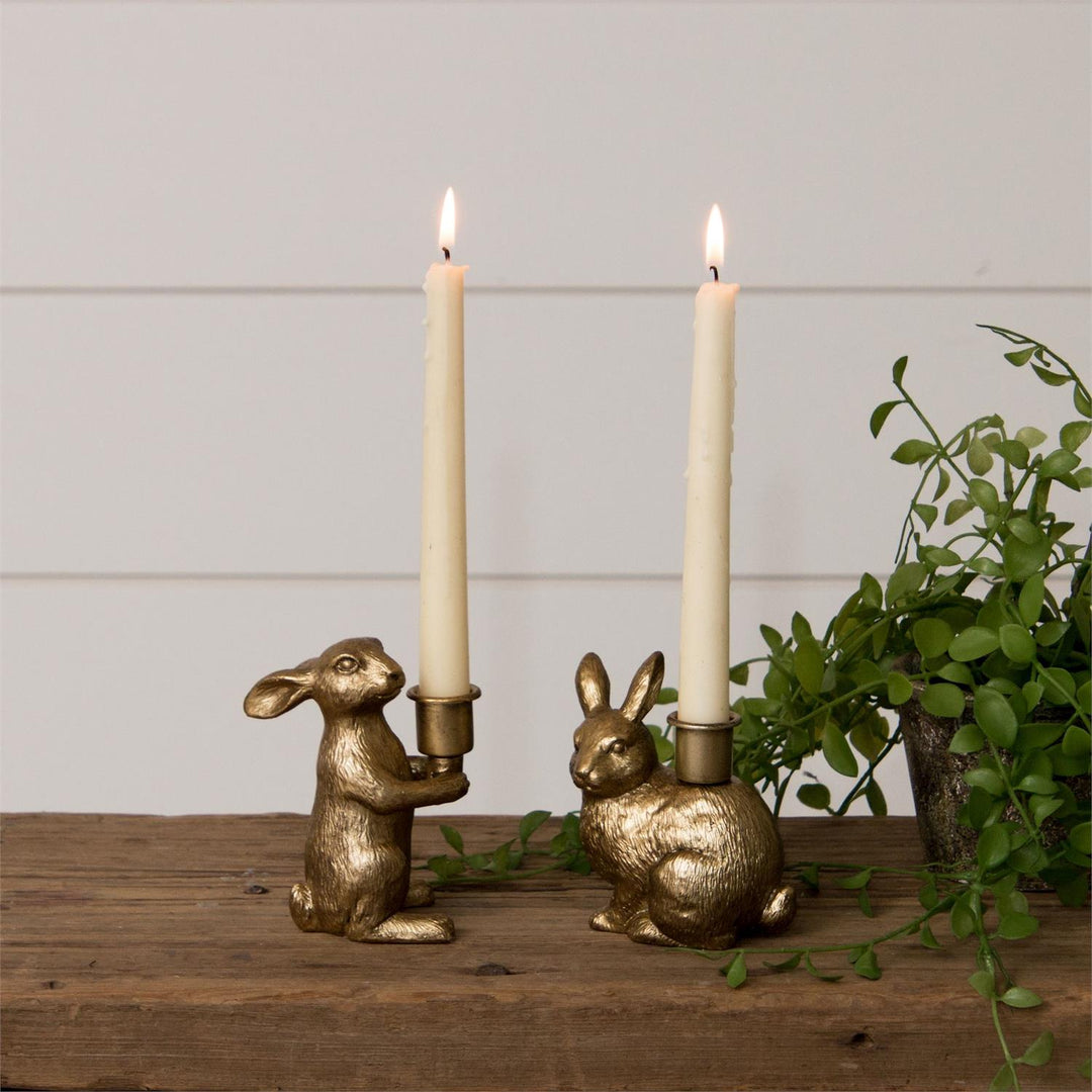 Two gold rabbit-shaped candle holders with lit candles on a wooden surface with a plant in the background.