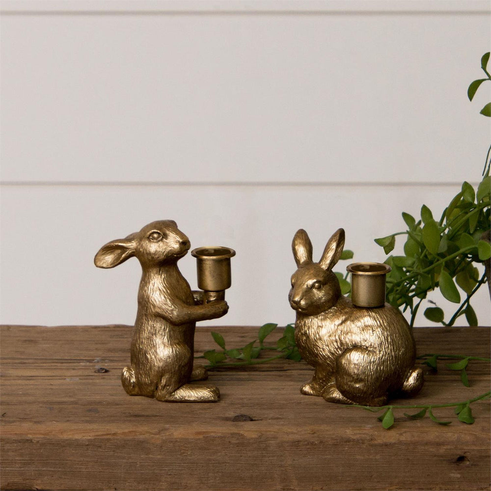 Two gold rabbit-shaped candle holders on a wooden surface with a white wall and plant in the background.