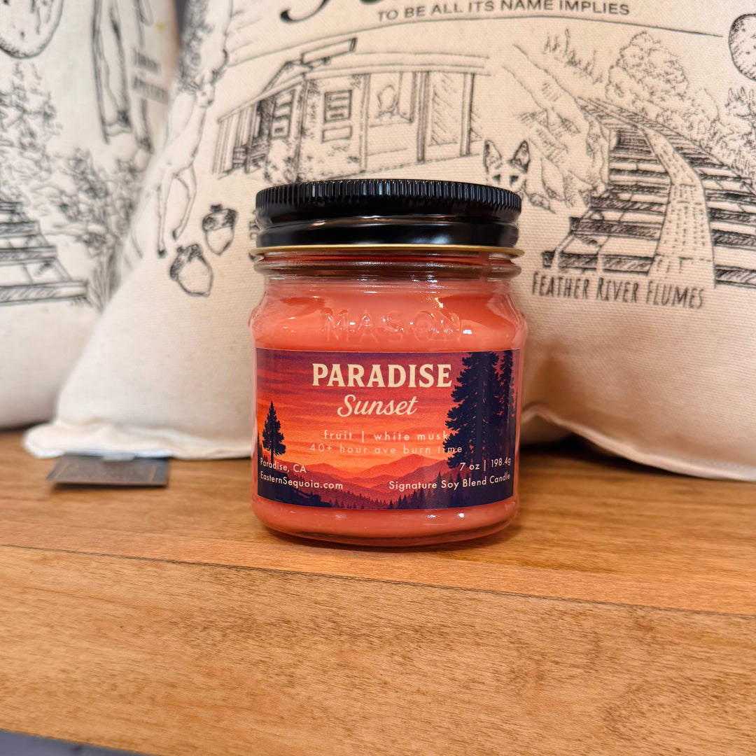 Jar of 'Paradise Sunset' candle on a wooden surface with a decorative pillow in the background.