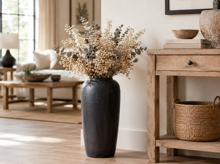 A tall matte black ceramic vase filled with dried beige and gray botanical stems, styled in a bright, modern living room next to a rustic wooden console table.