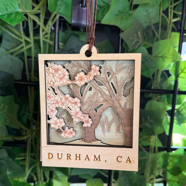 Wooden hanging ornament with floral design and 'Durham, CA' text, against a green leafy background.