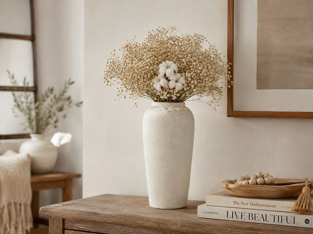A tall textured off-white ceramic vase filled with dried baby's breath and cotton stems, styled on a wooden console table in a warm, neutral-toned room.