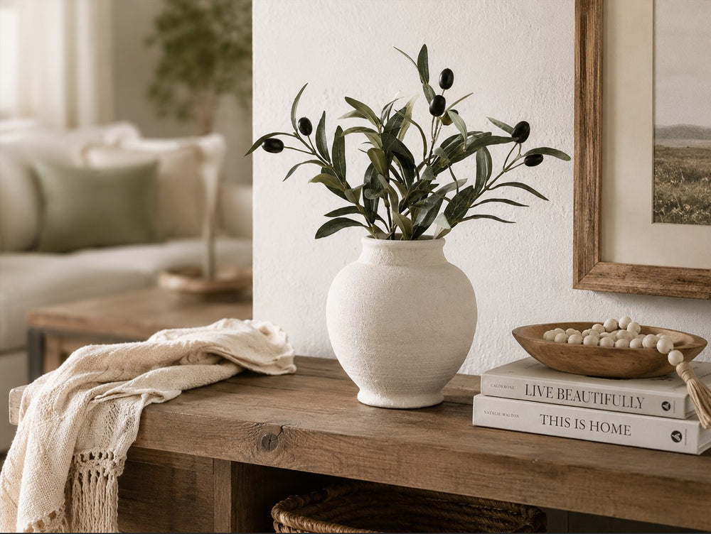 A textured off-white ceramic vase filled with green olive branches, styled on a rustic wooden console table next to books and a wooden bowl of white beads in a warm, modern living room setting.