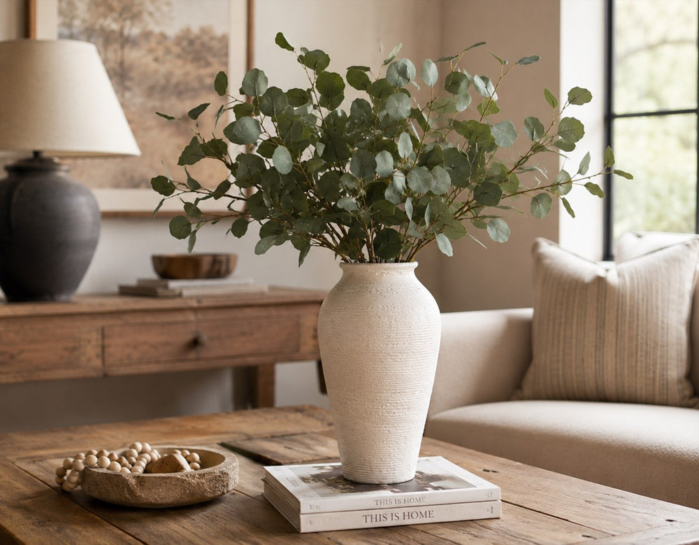 Textured white ceramic farmhouse vase with eucalyptus branches styled on a rustic wood coffee table in a warm neutral living room.