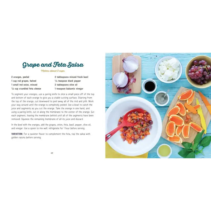 A printed recipe card for Grape and Feta Salsa shown beside a styled food preparation scene. The left side contains the recipe text, and the right side shows oranges, grapes, garlic, bowls of ingredients, and a plated salsa with garnish on a light blue surface.