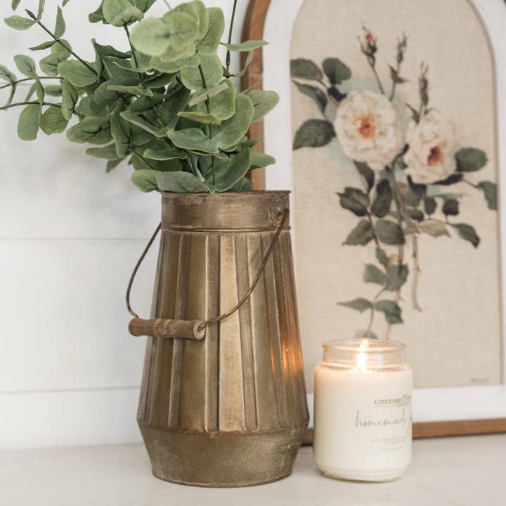 Bronze vase with vertical ribbing and a tall swing-style handle, displayed with green foliage beside floral artwork