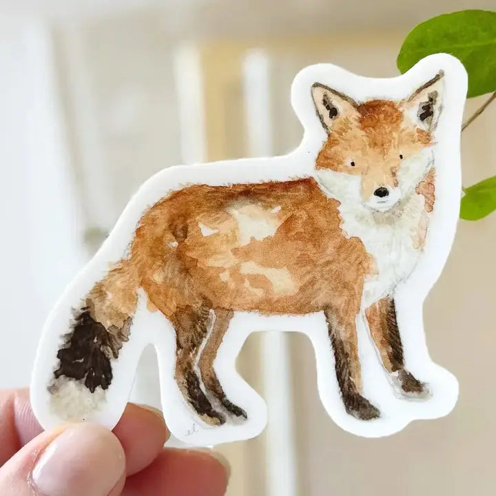 A die-cut sticker featuring a watercolor-style illustration of a fox in orange, brown, white, and black, held by a person&