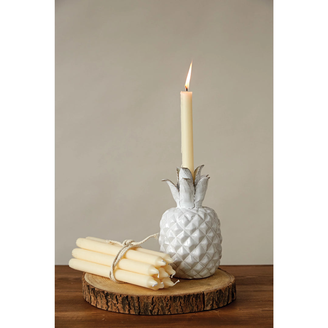 White pineapple-shaped candle holder with a lit candle and bundle of candles on a wooden surface.