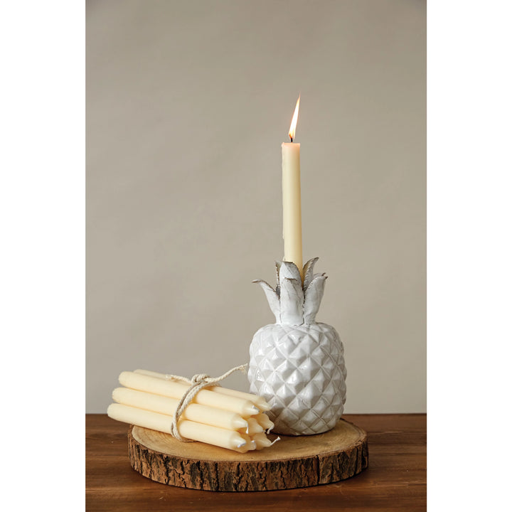 White pineapple-shaped candle holder with a lit candle and bundle of candles on a wooden surface.