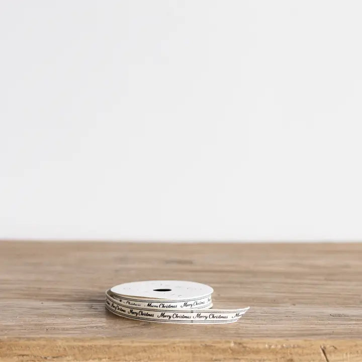 Narrow cream Christmas ribbon spool printed with black cursive Merry Christmas lettering on a wood tabletop.