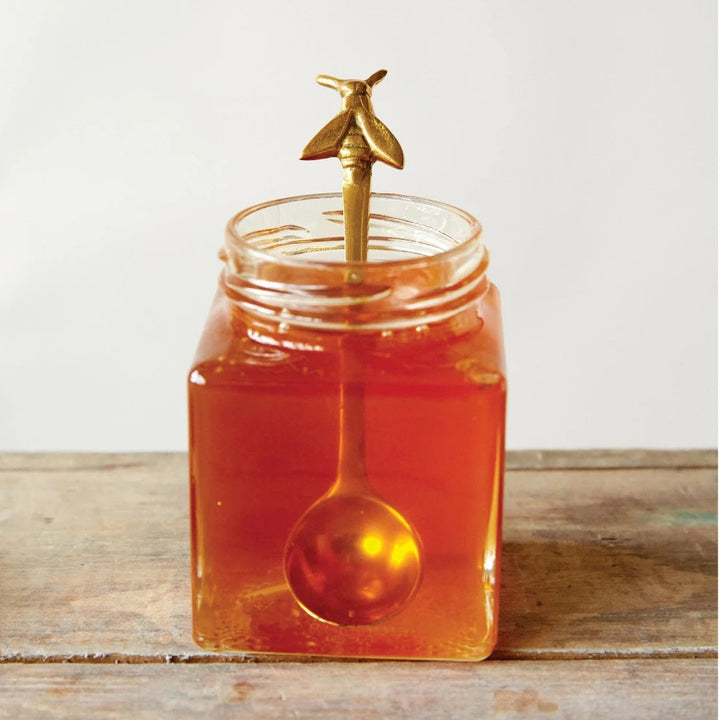 Jar of honey with a gold spoon and honey dipper on a wooden surface