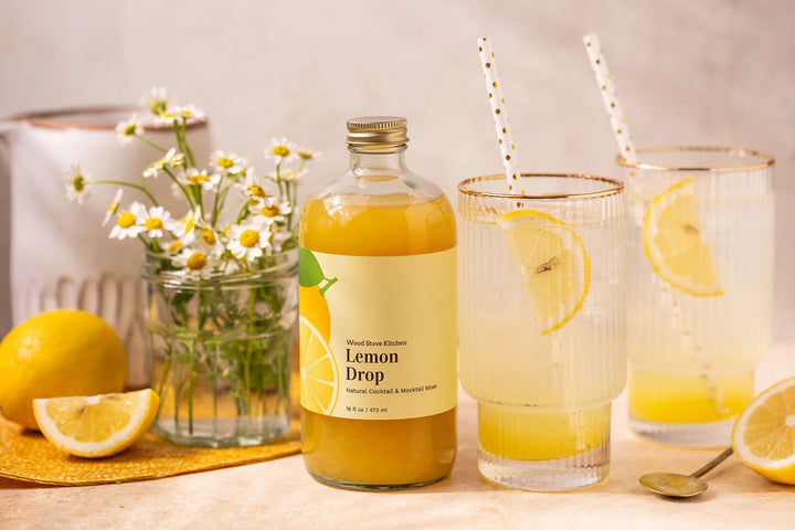 Bottle of Lemon Drop juice with two glasses and lemons on a table.