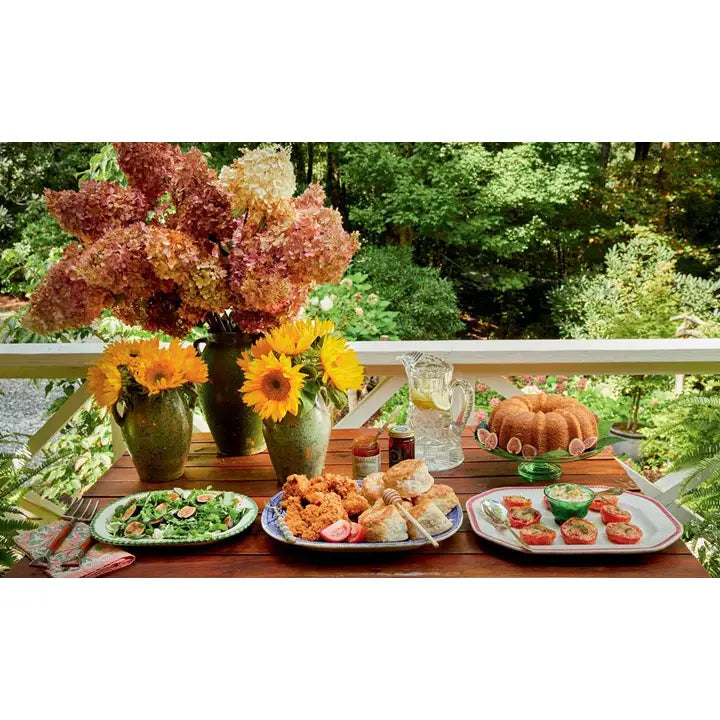 A styled outdoor dining table on a porch with a lush green garden background, featuring a large floral arrangement, sunflowers, a bundt cake, and plates of food including a salad, fried appetizers, and tomato and cheese skewers.