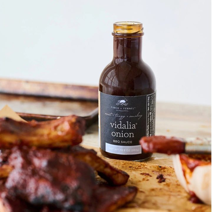Bottle of 'Vidalia Onion' BBQ Sauce on a wooden surface with ribs and a brush.