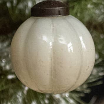 Ivory round glass Christmas ornament with vertical ribbing and a dark metal-look cap among evergreen branches