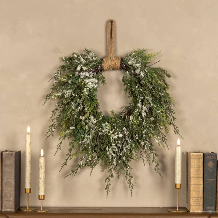 Snowy green cedar-style wreath with white snow accents and a rope hanging loop against a warm neutral wall