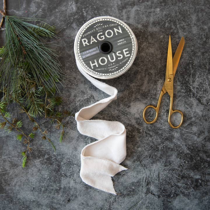 White velvet wired ribbon unrolled from a spool beside evergreen sprigs and gold-colored scissors